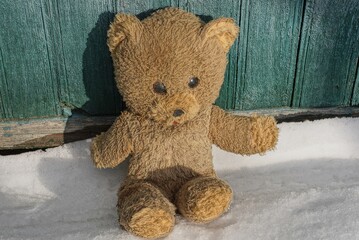 one old plush toy bear on a white snowdrift near a green wooden wall on a winter street