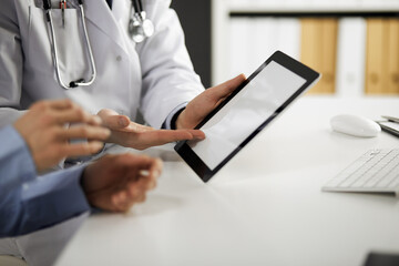 Unknown male doctor and patient woman discussing current health examination while sitting in clinic and using tablet computer, closeup of hands. Medicine and healthcare concept