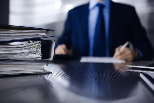 Binders with papers are waiting to be processed by man accountant in blue blazer staying at home during covid pandemic. Taxes and audit concept. Internal Revenue Service inspector checking financial