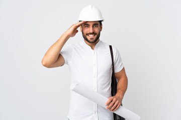 Young architect man with helmet and holding blueprints isolated on white background saluting with hand with happy expression
