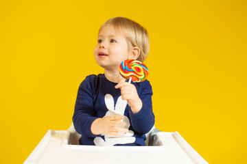 Cute little boy holding colorful lollipop while against sitting on chair over yellow background.