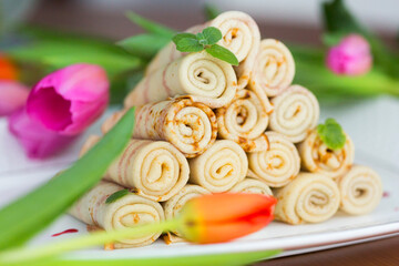Traditional Russian blini or pancakes for Maslenitsa or pancake week wrapped in rolls and stacked in a pyramid and pink tulips