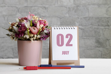 July 02. 02th day of the month, calendar date. Delicate bouquet of flowers in a pink vase, two pencils and a calendar with a date for the day on a wooden surface
