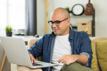 Indian man using laptop on sofa in living room texting on laptop sending message or chatting with online social media.