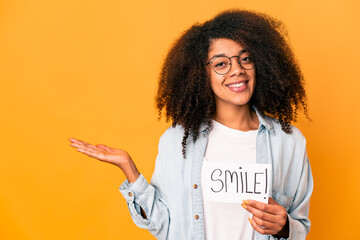 Young african american curly woman holding a smile message placard showing a copy space on a palm and holding another hand on waist.