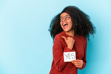 Young african american curly woman holding a yes placard points with thumb finger away, laughing...