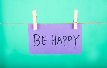 Be Happy word written on a Purple color sticky note hanging with a wire in a Cyan background.