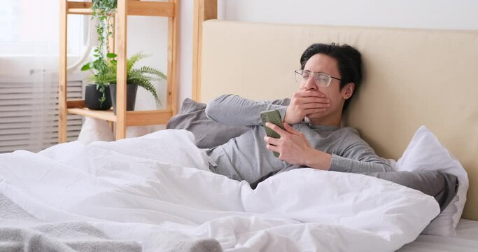 Man yawning and stretching on bed while using mobile phone at home