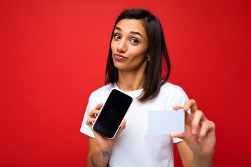 Portrait photo of self-confident brunette young woman wearing everyday stylish white t-shirt isolated on red background wall holding and using phone with empty screen for mockup and credit card making
