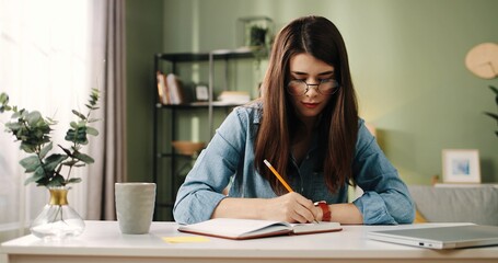 Young beautiful student girl with glasses sitting at desk in apartment and writes into the diary task for week.