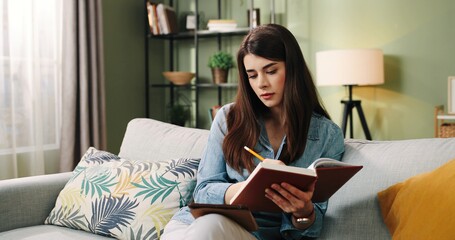 Naklejka premium Beautiful caucasian girl sitting at home on couch and writing in notebook lecture concept looking at tablet.