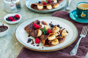 Thin chocolate pancakes stuffed with bananas, raspberries and blueberries, poured with chocolate sauce, on a gray ceramic plate on a light concrete background. Pancake recipes.