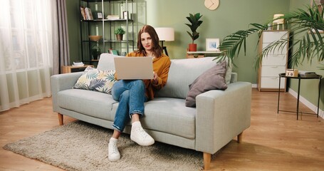 Caucasian beautiful happy young woman resting on couch in living room in house and surfing internet on laptop. Female worker working on computer at home typing and browsing. Freelance concept