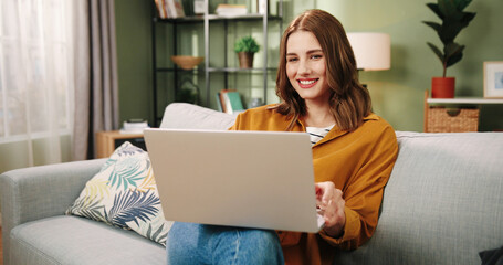 Portrait of happy joyful smiling beautiful Caucasian young female speaking on online video call on internet on laptop waving hand while sitting in cozy living room on sofa at home. Video chat concept