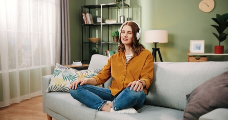 Young beautiful caucasian girl sitting at home alone on couch in headphones and enjoys music singing song.
