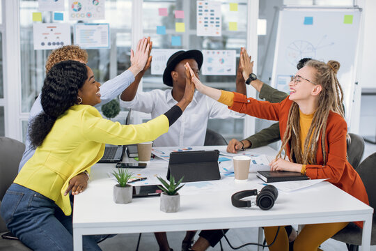 Multicultural casually dressed freelancers giving high five after finishing working meeting at office. Five colleagues feeling excited about start of new project.