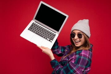 Side profile Photo shot of laughing crazy beautiful smiling dark blond young woman holding computer laptop with empty monitor screen with mock up and copy space wearing sun glasses hat and colourful