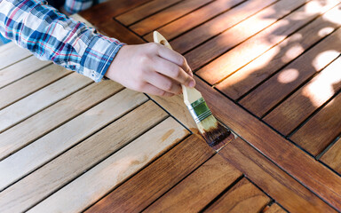 hand holding a brush applying varnish paint on a wooden garden table - painting and caring for wood with oil - 418946432