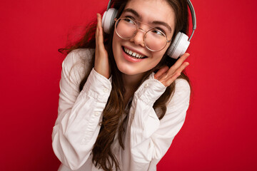 Closeup photo of attractive positive smiling young brunet woman wearing white shirt and optical glasses isolated over red background wearing white wireless bluetooth headsets listening to music and