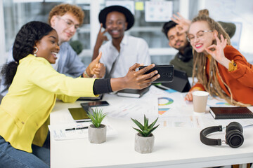 Blurred view of smiling creative multiracial colleagues, gesturing and laughing, while making selfie photo at the meeting in office room. Focus on hand of african woman, making photo