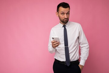 Thoughtful handsome young man isolated over background wall wearing everyday clothes holding and using mobile phone writing sms looking at camera and thinking