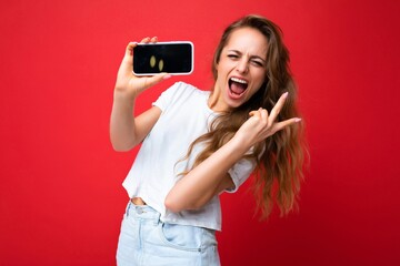 joyful smiling young blonde woman good looking wearing white t-shirt standing isolated on red background with copy space holding phone showing smartphone in hand with empty screen display for mockup