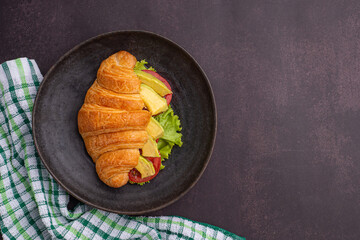 Top view of a croissant with avocados sliced, tomato, and lettuce on a black plate placed on a dark gray background. Space for text. Concept of foods