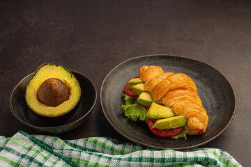 Croissant with avocado sliced, tomato, and lettuce, and avocado half on a black plate placed on a dark gray background. Space for text. Close-up photo. Concept of healthy foods and fruits