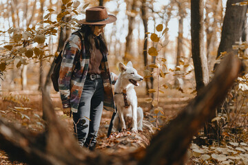 Obraz na płótnie Canvas Stylish woman with backpack hiking with white dog in sunny autumn woods. Cute swiss shepherd puppy