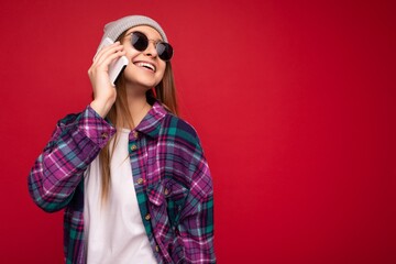 Closeup photo of charming smiling positive young woman wearing hipster purple shirt and casual white t-shirt grey hat and sunglasses isolated over red background holding in hand and talking on mobile