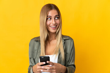 Young blonde woman isolated on yellow background using mobile phone and looking up