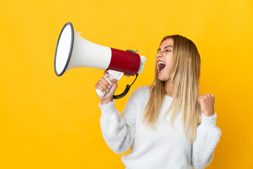 Young blonde woman isolated on yellow background shouting through a megaphone to announce something...
