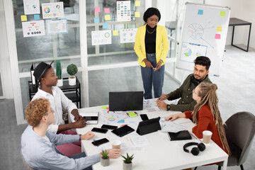 Office work concept. Pretty young African lady discussing strategy of joint project with her creative successful motivated diverse team, using infographics and gadgets in office room