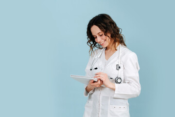 Charming female nurse writing in a notebook and smiling. Over blue background.