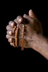 Praying hand of an old Indian Catholic man with wooden rosary isolated on a plain black background.