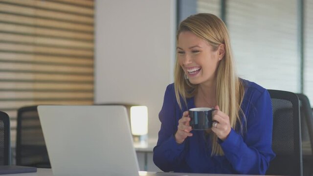Young businesswoman holding cup of coffee having late night video chat sitting at desk with laptop in modern open plan office - shot in slow motion