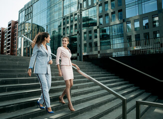 Two smiling business women talking while walking down stairs near modern office building. © Raisa Kanareva