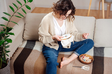 Peaceful young woman sewing in comforts of her apartment, embroiders a picture