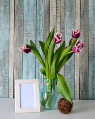 Spring still life with bouquet of lilac fringed tulips in glass bowl and blank white wooden frame on white table. Interior decorations. Natural light.  Vertical view 