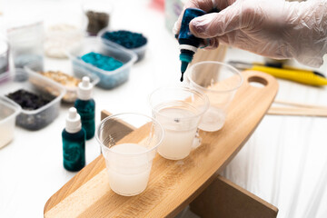 Epoxy resin DIY. A tube of blue dye in the artist's hand, against the background of three glasses with resin, standing on a wooden board