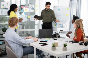 Group of mixed race startup team discussing creative ideas at office. Young multiracial colleagues sitting at table with charts, graphs and modern technology.
