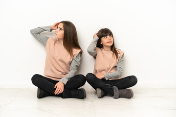 Little sisters sitting on the floor isolated on white background having doubts while scratching head