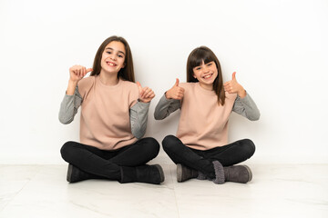 Fototapeta na wymiar Little sisters sitting on the floor isolated on white background giving a thumbs up gesture with both hands and smiling