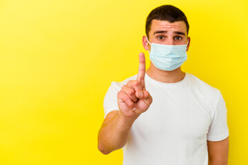 Young caucasian man wearing a protection for coronavirus isolated on yellow background showing number one with finger.