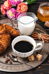 Croissant with chocolate hazelnut spread and cup of coffee on a metal vintage tray. Continental breakfast with flowers on dark wooden table