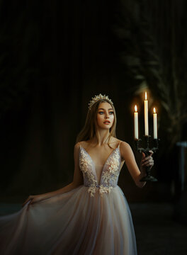 Art photo of medieval girl princess walks in dark gothic room. Woman queen is holding candlestick with burning candles in hand. Pink purple dress, long loose blonde hair gold royal crown. Fairy image