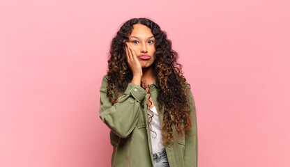 young hispanic woman feeling shocked and astonished holding face to hand in disbelief with mouth...
