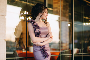 Fototapeta premium Closeup of luxury young woman leaning on modern sliding restaurant window and posing for camera in the evening