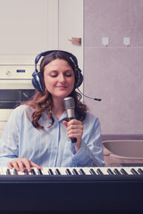 Woman with headphones sings a song into the microphone and plays the synthesizer in the home kitchen