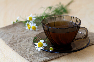 Fototapeta na wymiar dark glass cup of Herbal tea with chamomile tea on sackcloth on a light wooden table with copy space. cup fo tea with bouquet fresh flowers and green leaves on yellow background
